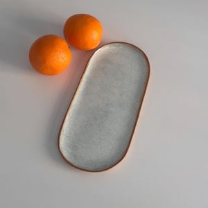 May include: A white oval ceramic serving tray with a speckled glaze. The tray is sitting on a white surface with two orange fruits beside it.