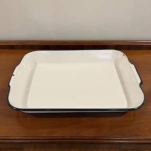 May include: A rectangular, cream-coloured enamel baking tray with a black trim. The tray has gently curved sides and small handles. The interior is smooth and reflective, suggesting it's been well-used.