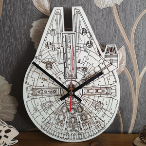 May include: A white wooden clock shaped like the Millennium Falcon from Star Wars. The clock face is detailed with the ship's interior and has black hands.
