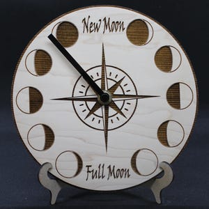 May include: A wooden clock with a compass design and moon phases around the edge. The clock face reads "New Moon" at the top and "Full Moon" at the bottom.