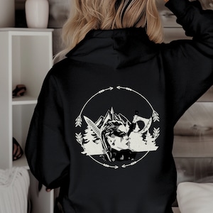 May include: Black hooded sweatshirt with a white graphic print of a woman with an axe in a forest scene. The graphic is surrounded by a circle with arrows.