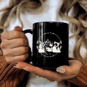 May include: Black ceramic mug featuring a white graphic of a warrior with mountains, trees, and a sword inside a circular frame. The mug has a black handle and is held by a person.