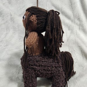 May include: A handmade, brown and tan crocheted centaur doll. The doll has a human upper body with dark brown hair in a ponytail and a horse-like lower body. The doll is made of yarn and has a whimsical appearance.