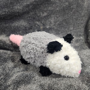May include: A plush toy shaped like an opossum with a grey, white, and black fur pattern. The opossum has a pink nose and black eyes.