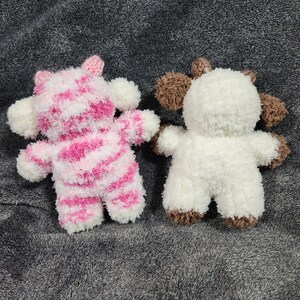 Puede incluir: Dos animales de peluche, uno de rayas rosas y blancas y otro blanco con detalles marrones. Los animales est&aacute;n sentados sobre una superficie gris y peluda.