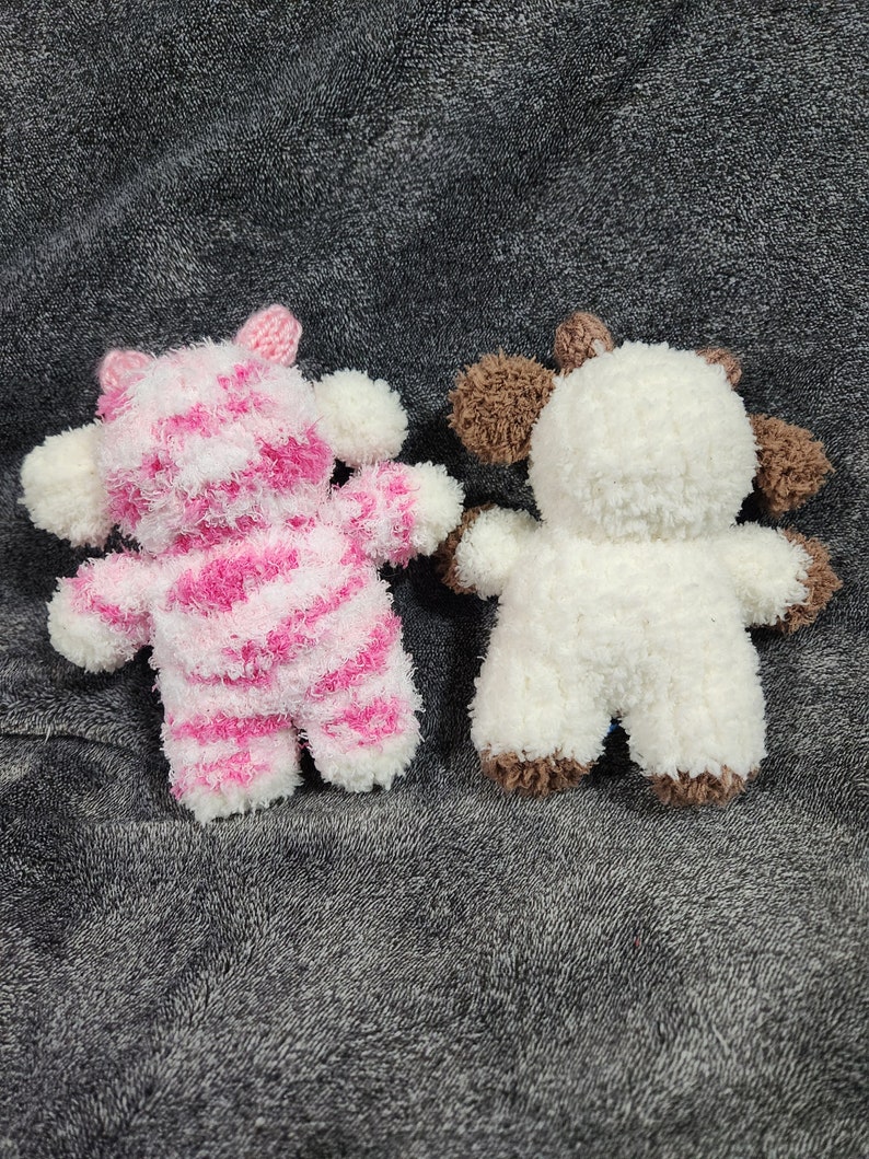 Puede incluir: Dos animales de peluche, uno de rayas rosas y blancas y otro blanco con detalles marrones. Los animales est&aacute;n sentados sobre una superficie gris y peluda.