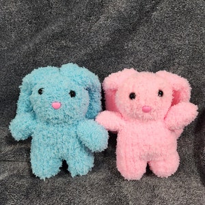 May include: Two plush bunny toys, one blue and one pink, with black eyes and pink noses. The toys are sitting on a gray fuzzy surface.