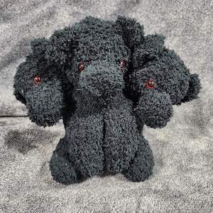 May include: A black plush toy with three heads and red eyes. The toy is sitting on a grey surface.