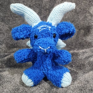 Puede incluir: Un juguete de peluche azul tejido en forma de cabra con cuernos azul claro. El juguete tiene una textura esponjosa y una expresión amigable.