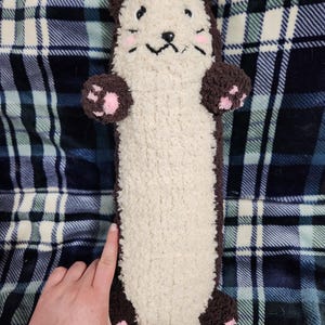 May include: A plush, long otter-shaped pillow with a cream-colored body and brown accents. The otter has a smiling face, pink cheek markings, and pink paw pads. The pillow is on a blue and white plaid blanket.