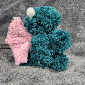 May include: A teal blue plush toy with a pink wing and a white pom-pom on its head. The toy has black eyes and a fuzzy texture.