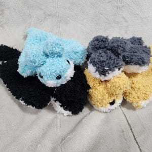 May include: A selection of handmade fox-shaped plush toys. One toy is light blue and black, the other is grey and yellow. They have white accents and black eyes. The toys are made from a soft, textured material.