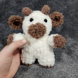 Puede incluir: Un animal de peluche blanco tejido a crochet con orejas marrones, una nariz marr&oacute;n y ojos negros. El animal tiene forma de vaca.