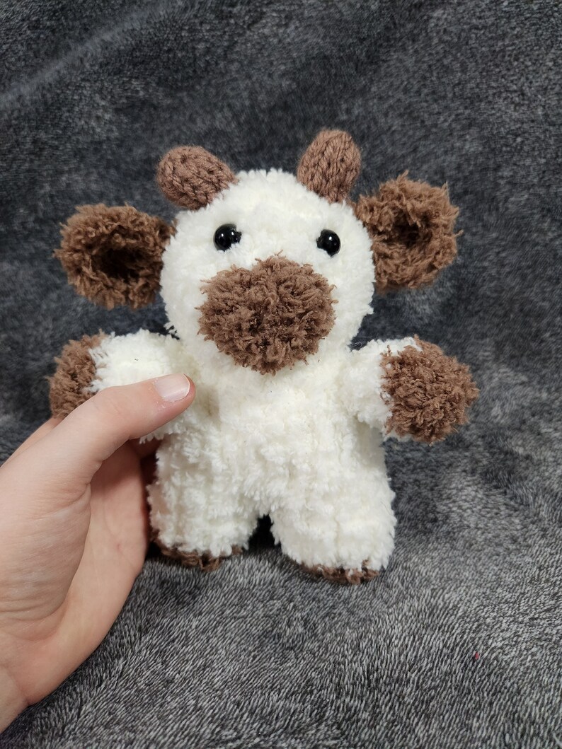 Puede incluir: Un animal de peluche blanco tejido a crochet con orejas marrones, una nariz marr&oacute;n y ojos negros. El animal tiene forma de vaca.
