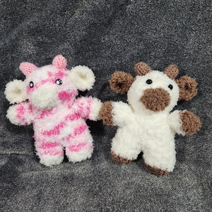 May include: Two crocheted stuffed cows, one pink and white striped and one white with brown accents. The cows are sitting on a gray fuzzy surface.