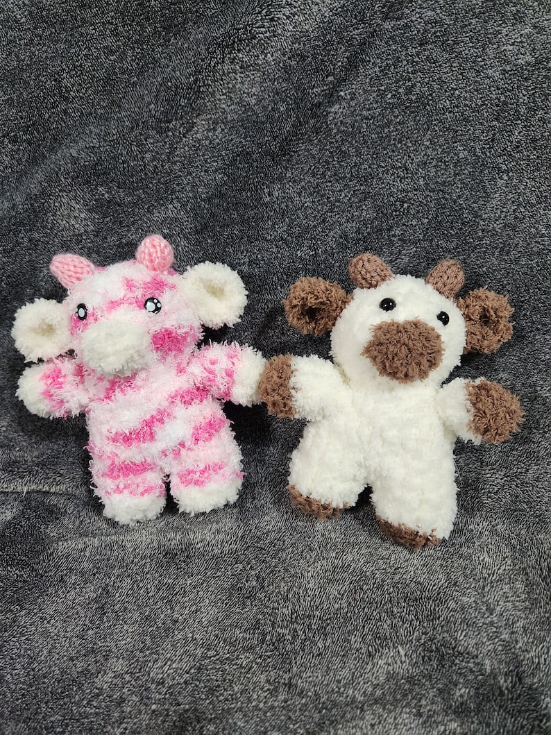Puede incluir: Dos vacas de peluche tejidas a crochet, una de rayas rosa y blancas y otra blanca con detalles marrones. Las vacas est&aacute;n sentadas sobre una superficie gris y peluda.