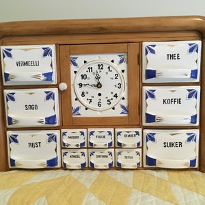 May include: A wooden spice rack with a clock face and 12 drawers. The drawers are labeled with Dutch words for various spices, including "Vermicelli", "Sago", "Thee", "Koffie", "Rijst", and "Suiker".
