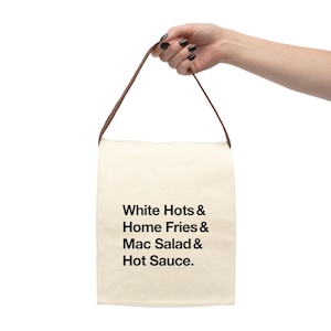 May include: A cream-colored tote bag with a brown handle is held up against a white background. The bag features black text that reads "White Hots & Home Fries & Mac Salad & Hot Sauce."