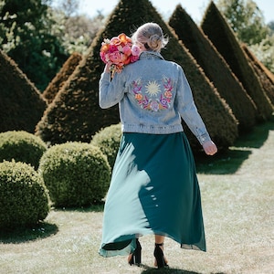 Pode incluir: Casaco de ganga claro com bordados florais coloridos, combinado com uma saia longa verde-azulado. A pessoa segura um buquê de flores rosa e laranja. O fundo apresenta sebes cuidadas e um ambiente ensolarado ao ar livre.