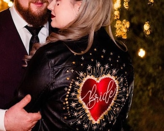 May include: A couple poses for a wedding photo. The groom wears a white shirt and a burgundy vest. The bride wears a black leather jacket with a red heart and the word "Bride" embroidered on the back.
