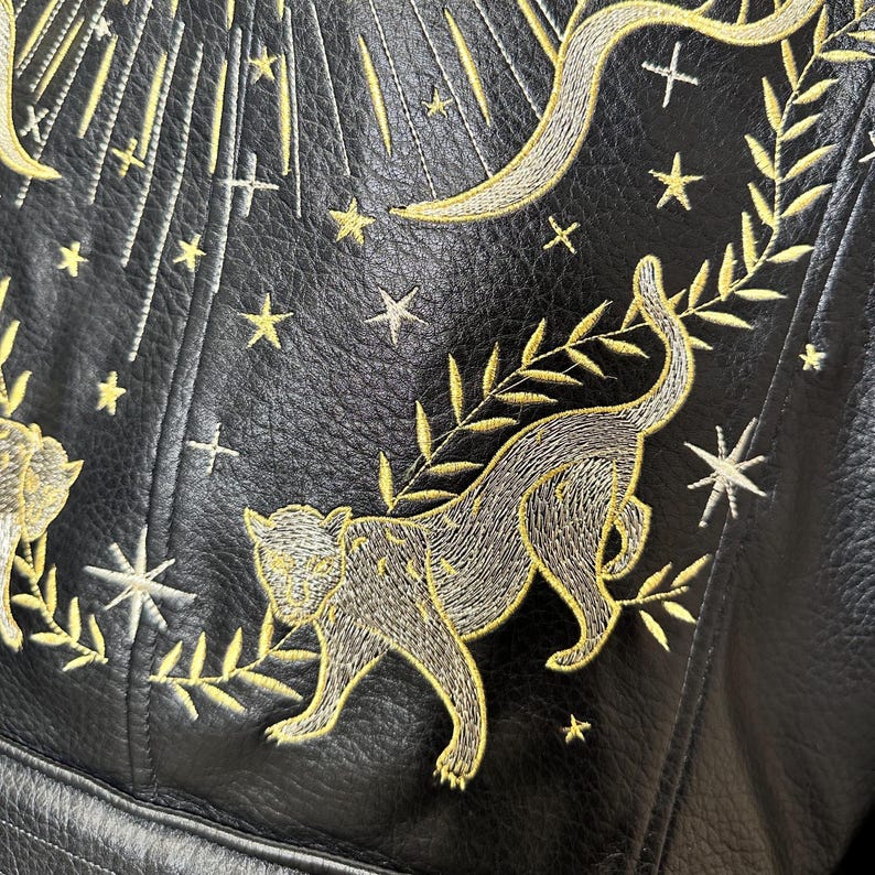 May include: Black leather jacket with intricate gold and silver embroidery. The design features a stylized panther, stars, and celestial elements. The stitching is detailed, creating a textured surface. The jacket's design is a blend of artistry and craftsmanship.