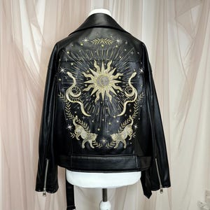 May include: Black faux leather jacket with gold embroidered celestial design on the back. The design includes a sun, snakes, stars, and two stylized cat figures. The jacket has a classic biker style with a notched collar and zipper details.