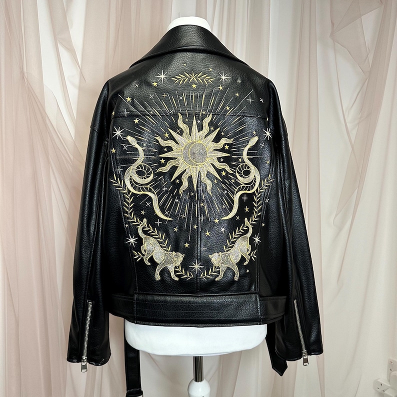 May include: Black faux leather jacket with gold embroidered celestial design on the back. The design includes a sun, snakes, stars, and two stylized cat figures. The jacket has a classic biker style with a notched collar and zipper details.