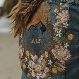 May include: Denim jacket with embroidered floral and mountain design. The back of the jacket features a mountain scene with trees, flowers, and the date 12.9.21 in gold thread. The jacket is a medium wash denim.