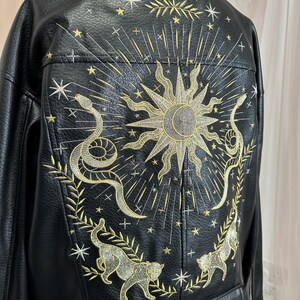 May include: Black faux leather jacket with gold embroidery. The back features a sunburst design, snakes, cats, and stars. The jacket has a classic collar and a tailored fit, perfect for adding a touch of celestial style to any outfit.