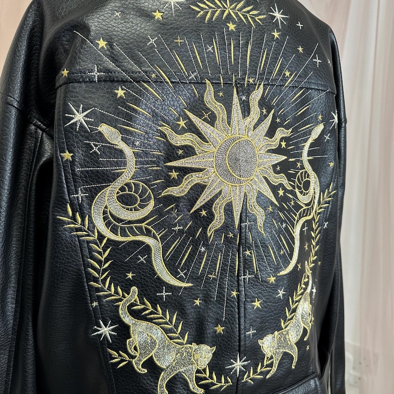 May include: Black faux leather jacket with gold embroidery. The back features a sunburst design, snakes, cats, and stars. The jacket has a classic collar and a tailored fit, perfect for adding a touch of celestial style to any outfit.