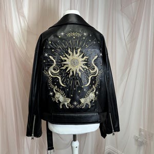 May include: Black faux leather jacket with a decorative embroidered design on the back. The design features a sun, snakes, stars, and two tigers in gold thread. The jacket has a notched collar, zipper details, and a belt.