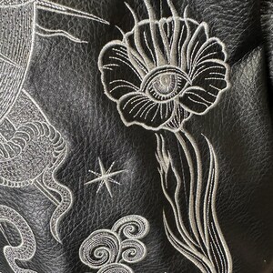 May include: Close-up of black leather with intricate white embroidery. The design features a large flower with an eye in the center, a star, and swirling cloud motifs. The stitching is detailed and creates a textured effect.