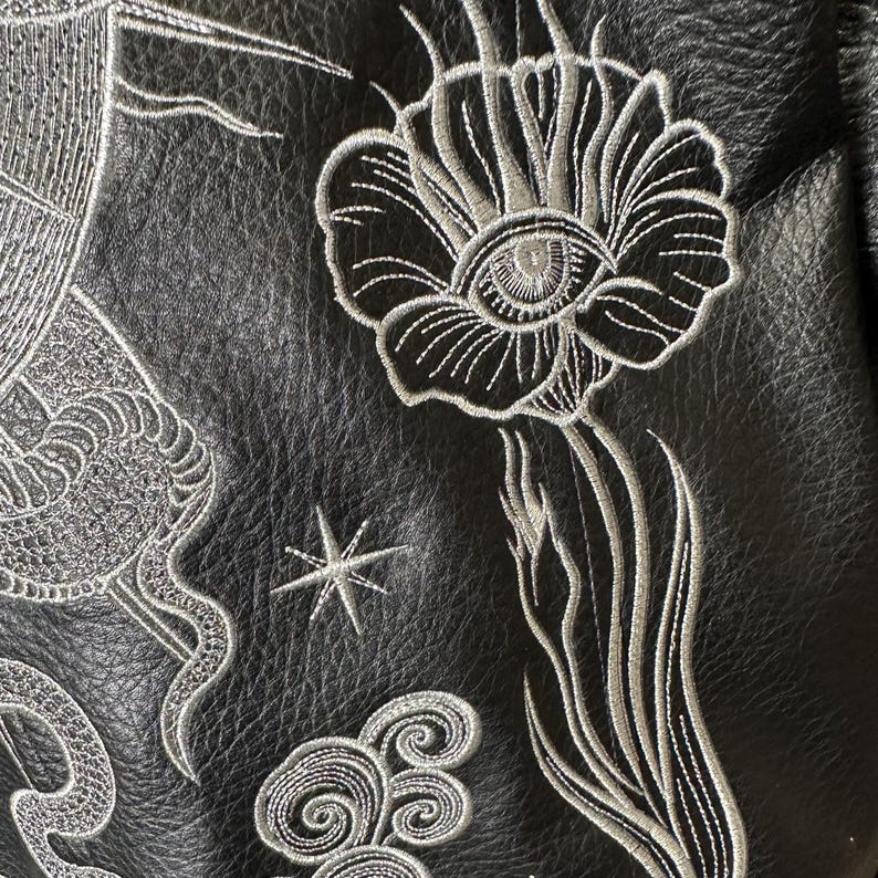 May include: Close-up of black leather with intricate white embroidery. The design features a large flower with an eye in the center, a star, and swirling cloud motifs. The stitching is detailed and creates a textured effect.