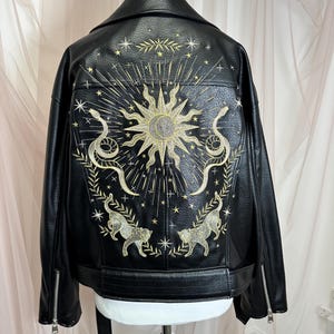 May include: Black faux leather jacket with gold embroidered sun, snake, and cat designs on the back. Features a classic biker jacket silhouette with a notched collar, zipper details, and a belt.