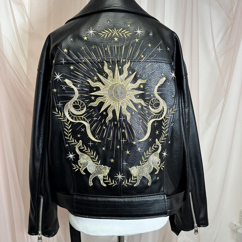 May include: Black faux leather jacket with gold embroidered sun, snake, and cat designs on the back. Features a classic biker jacket silhouette with a notched collar, zipper details, and a belt.