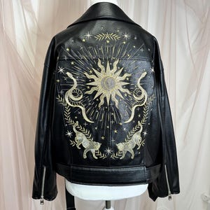May include: Black faux leather jacket with intricate gold embroidery on the back. The design features a sun, snakes, stars, and two cat-like figures. The jacket has a classic biker style with a notched collar and zipper details.