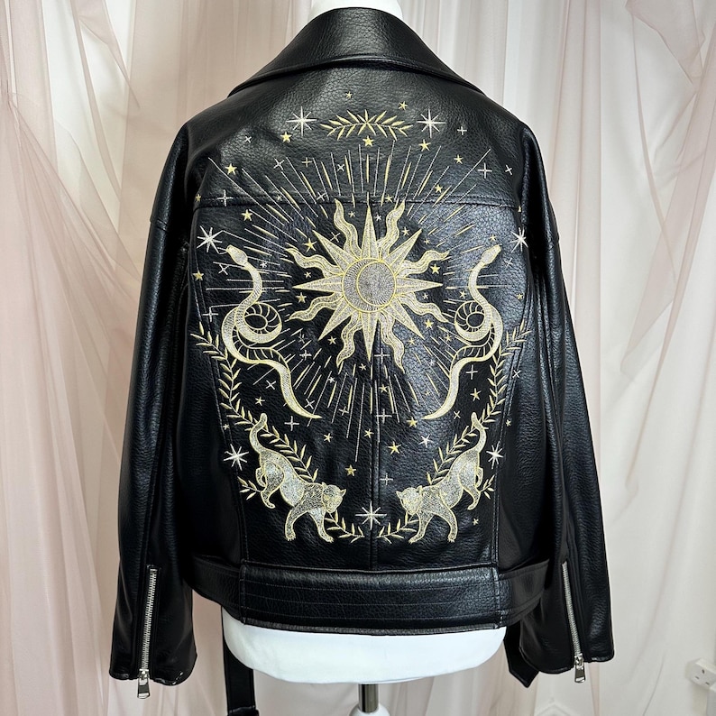 May include: Black faux leather jacket with intricate gold embroidery on the back. The design features a sun, snakes, stars, and two cat-like figures. The jacket has a classic biker style with a notched collar and zipper details.
