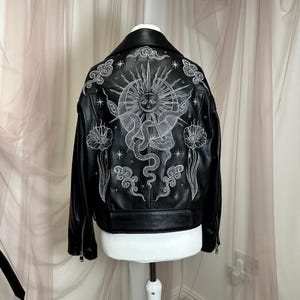May include: Black leather jacket with white celestial design on the back. The design includes a sun, snakes, clouds, and flowers. The jacket has a classic biker style with a notched collar and zipper details.