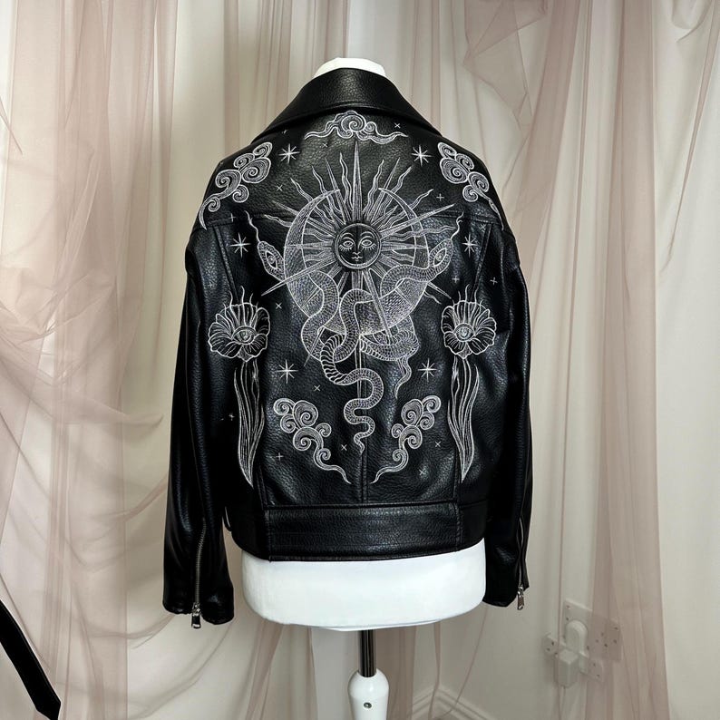 May include: Black leather jacket with white celestial design on the back. The design includes a sun, snakes, clouds, and flowers. The jacket has a classic biker style with a notched collar and zipper details.