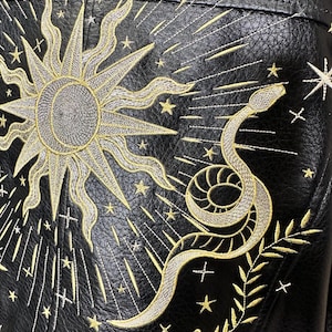 May include: Black leather jacket with gold embroidery featuring a sun, stars, and a snake. The sun has a detailed, textured center and radiating lines. The snake is coiled with intricate detailing, and the stars are scattered across the jacket.