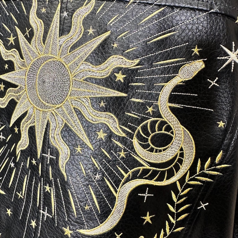 May include: Black leather jacket with gold embroidery featuring a sun, stars, and a snake. The sun has a detailed, textured center and radiating lines. The snake is coiled with intricate detailing, and the stars are scattered across the jacket.