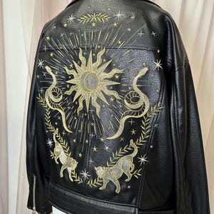 May include: Black faux leather jacket with intricate gold embroidery. The design features a sun, snakes, cats, and stars. The jacket has a classic biker style with a zipper closure.