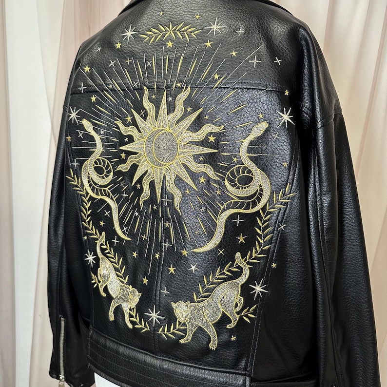 May include: Black faux leather jacket with intricate gold embroidery. The design features a sun, snakes, cats, and stars. The jacket has a classic biker style with a zipper closure.