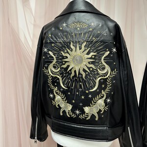 May include: Black faux leather jacket with gold embroidery on the back. The design features a sun, snakes, cats, stars, and celestial elements. The jacket has a classic biker style with a notched collar and zipper details.