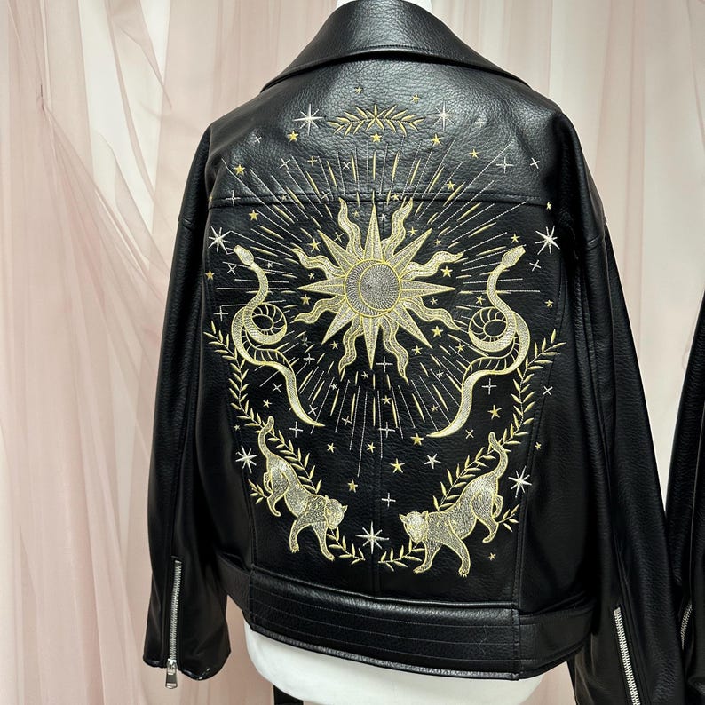 May include: Black faux leather jacket with gold embroidery on the back. The design features a sun, snakes, cats, stars, and celestial elements. The jacket has a classic biker style with a notched collar and zipper details.