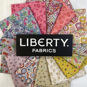 Puede incluir: Una colección de muestras de tela floral colorida dispuestas en un patrón circular. Las telas presentan varios diseños florales en tonos rosa, azul, amarillo y morado. Un letrero negro con el texto "LIBERTY. FABRICS" está en el centro.