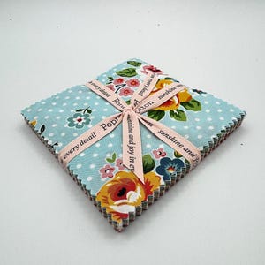 Puede incluir: Un conjunto de cuadrados de tela con lunares azul claro y motivos florales amarillos, rosas y azules. Una cinta rosa, con texto, asegura el conjunto. La cinta dice "sunshine and joy in every detail".