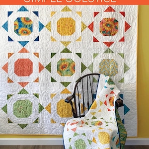 May include: A white quilt with colorful geometric patterns, including squares and triangles, hangs on a wall. The quilt features various colors and floral designs. The text "SIMPLE SOLSTICE" is displayed on an orange banner. A matching quilt is draped over a wooden chair.