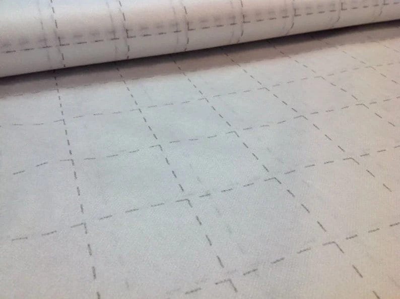 May include: A roll of white fabric with a grid pattern of dashed gray lines. The fabric is partially rolled, revealing the grid design across the surface. The pattern suggests a quilting or crafting material.