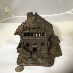 May include: A dark brown cast iron piggy bank shaped like a small house with a steeply pitched roof and a chimney. The house has a front door and windows, and the base is sculpted to look like a rocky hillside.